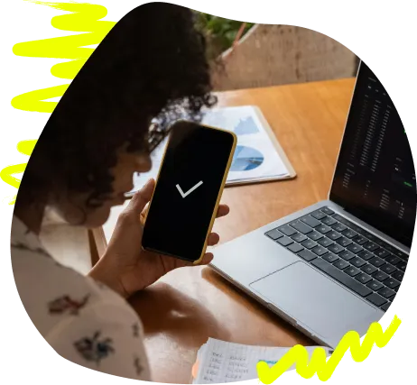 Person hält ein Smartphone mit Häkchen neben einem Laptop am Schreibtisch